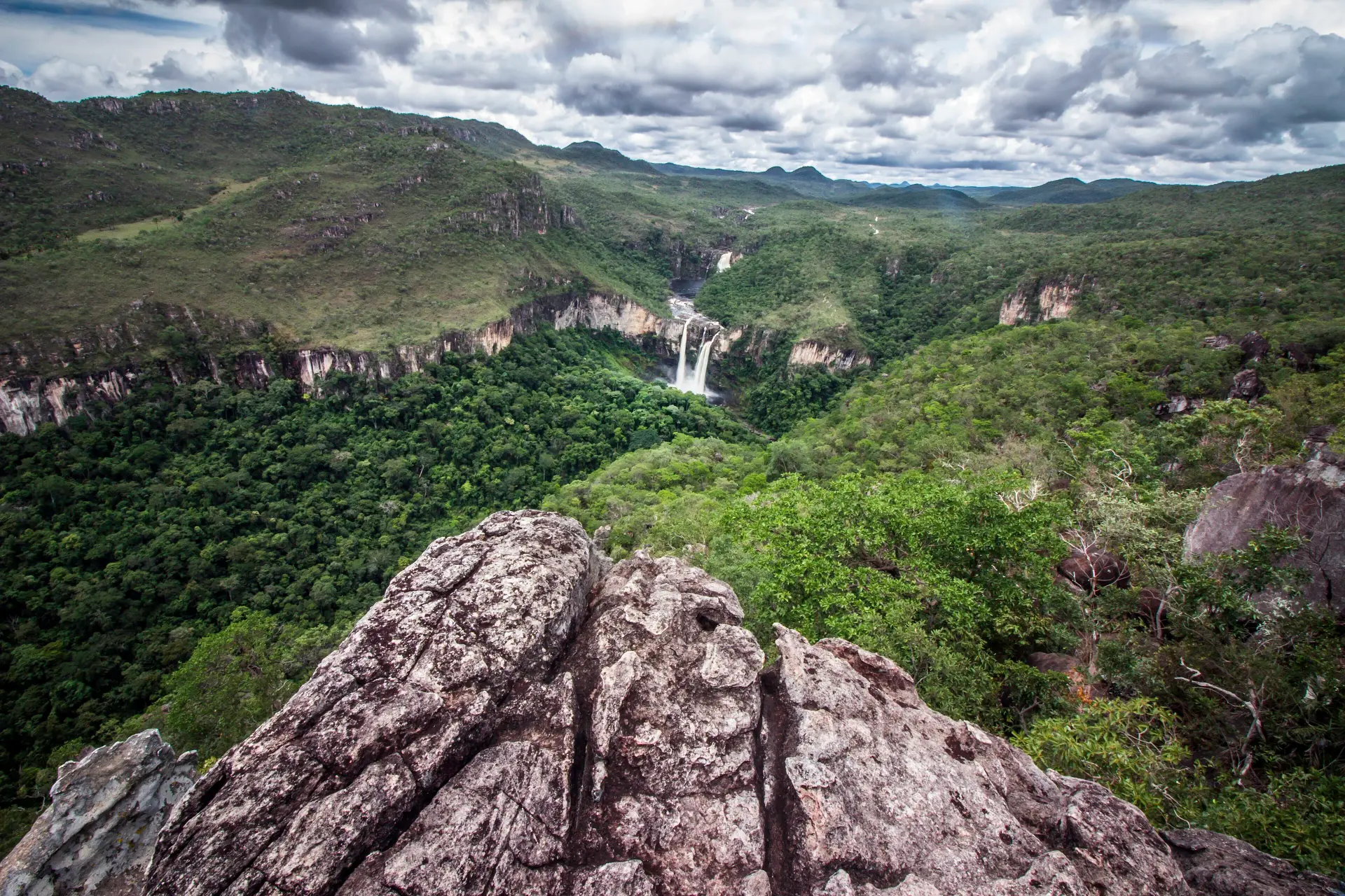 Green valley of the Chapada dos Veadeiros seen from a rock ledge, with a waterfall far below under a cloudy sky
