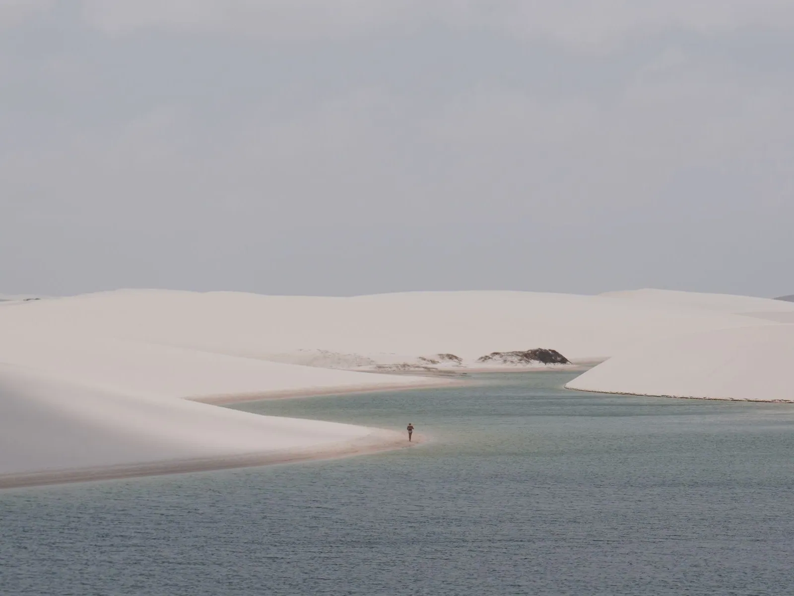 A single walker on a dune crest above a turquoise rainwater lagoon in the Lençóis Maranhenses