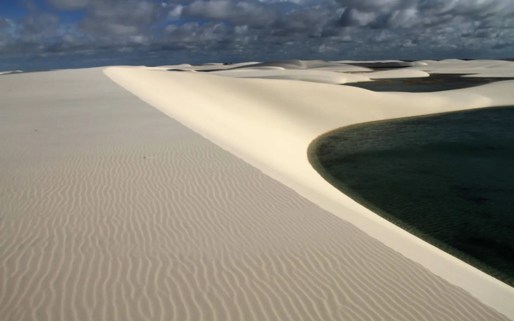 A rippled white dune sweeping down to the green water of Lagoa das Gaivotas under evening cloud