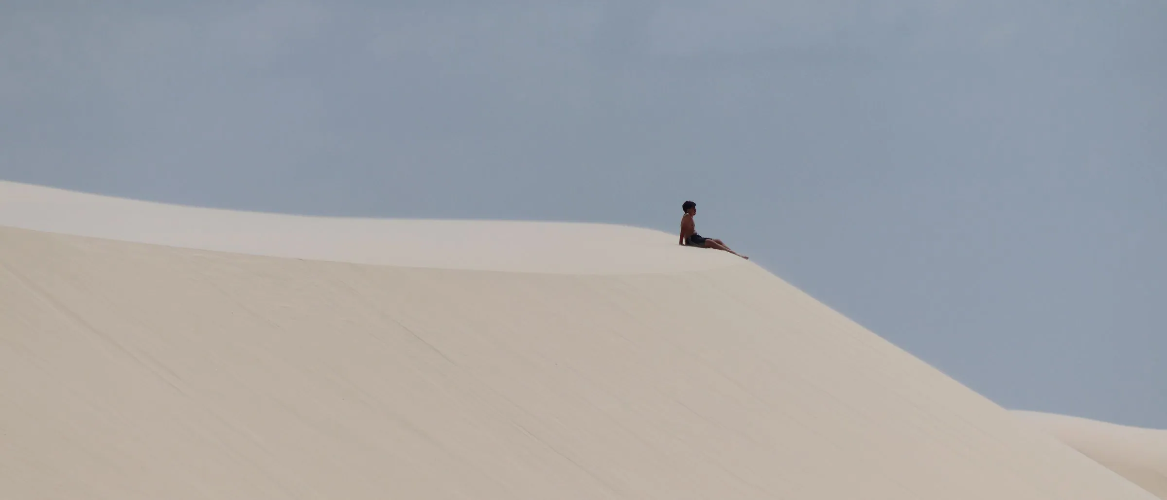 A lone figure sitting on a vast dune crest, the sand sea fading toward the horizon