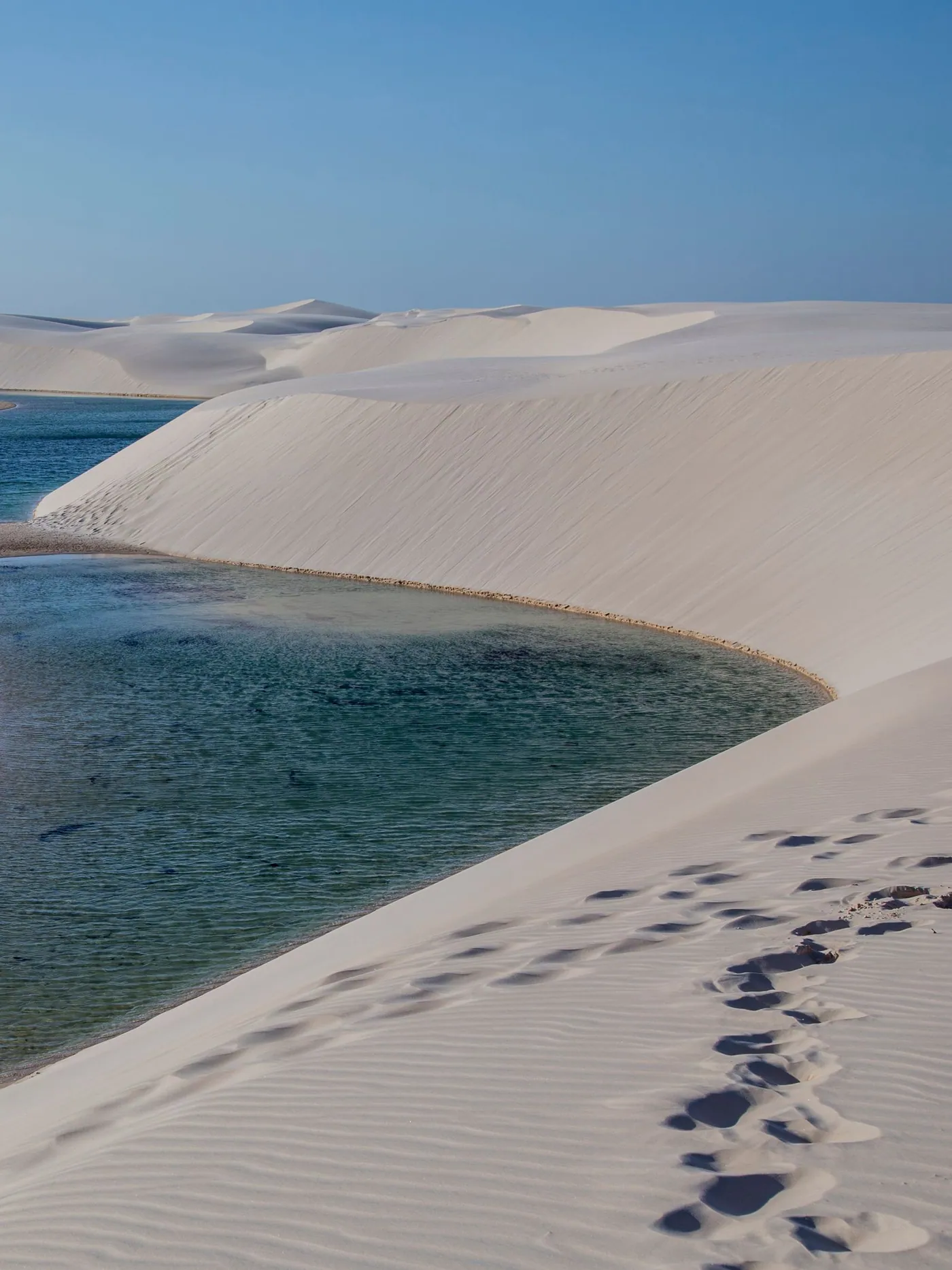 A single line of footprints crossing white sand to the edge of a still blue lagoon