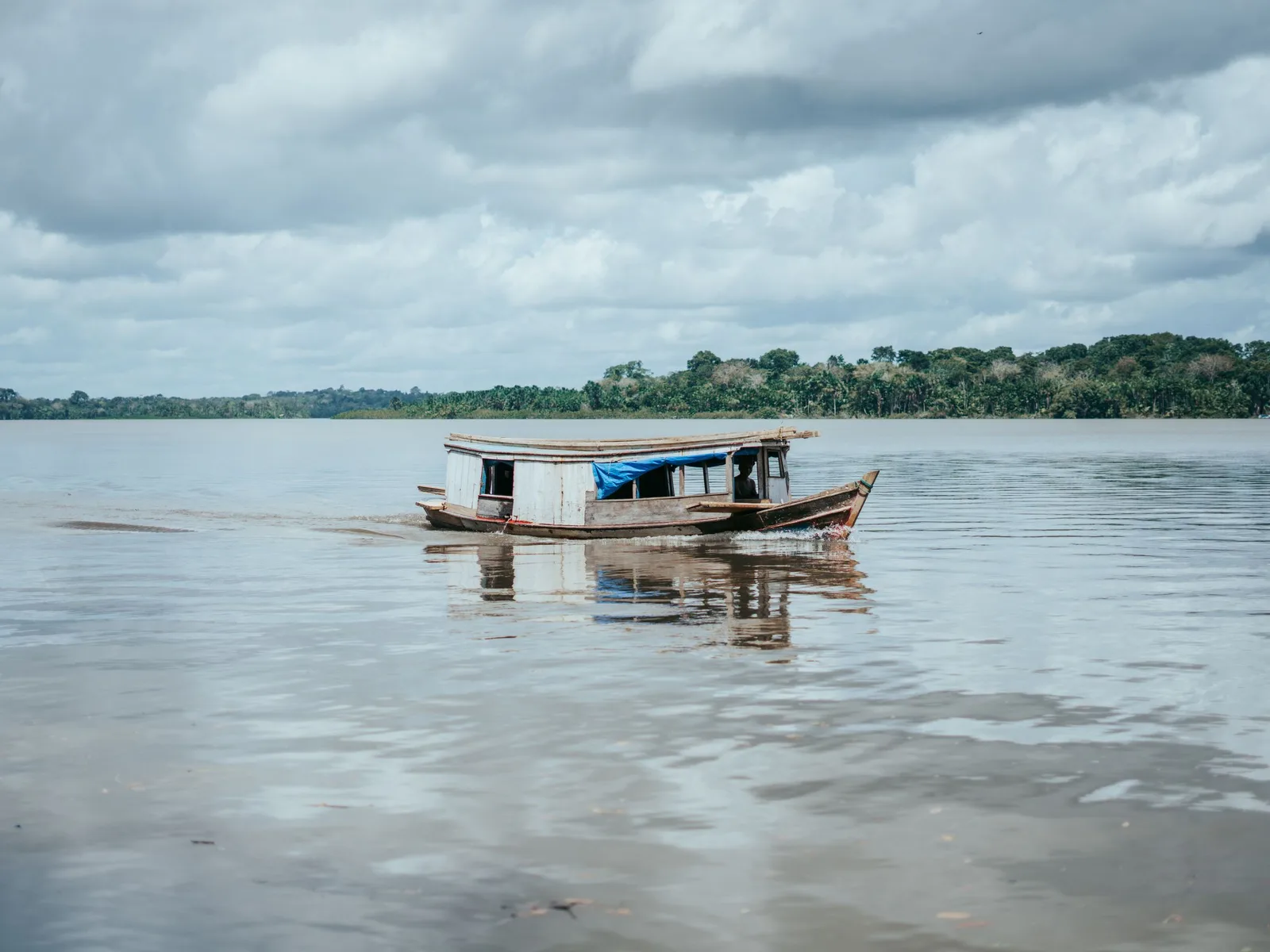 A small wooden passenger boat crossing a wide, flat river under heavy cloud