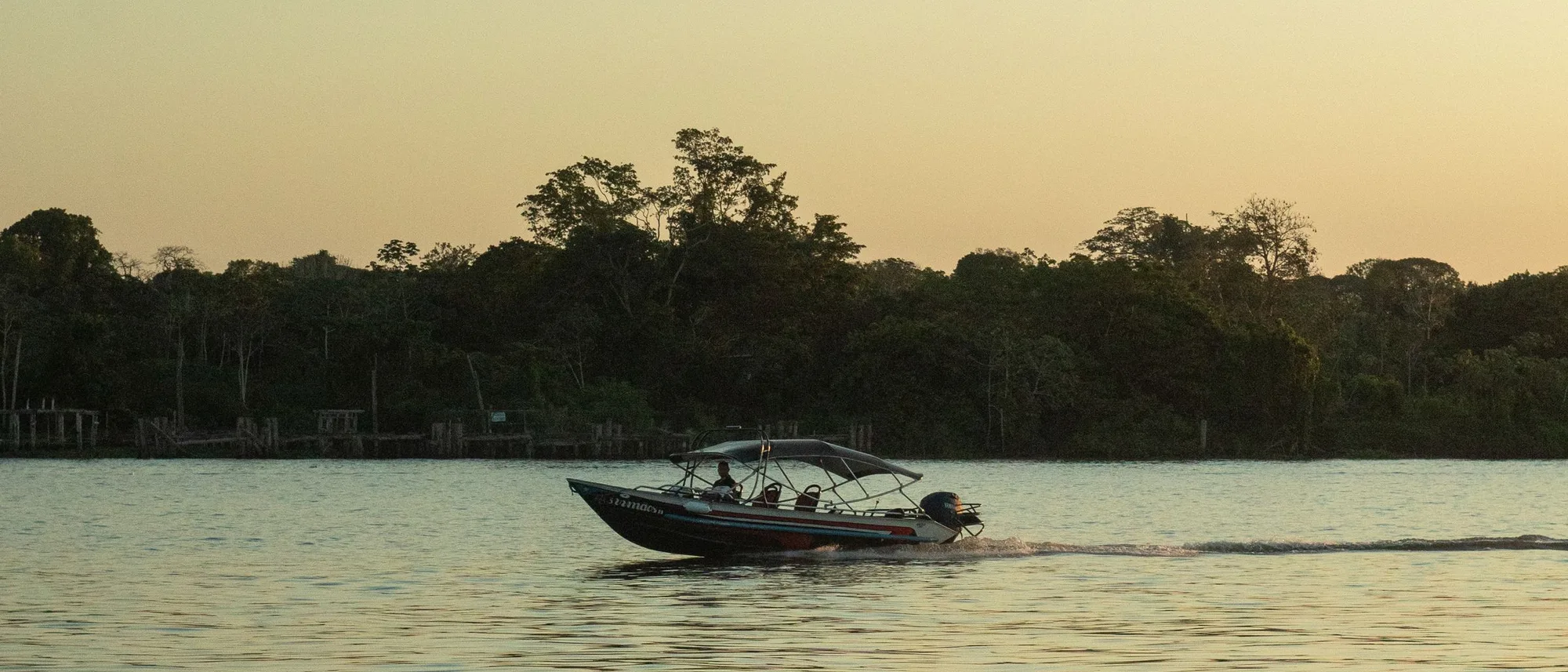 A small boat crossing wide flat water at dusk beneath a dark treeline
