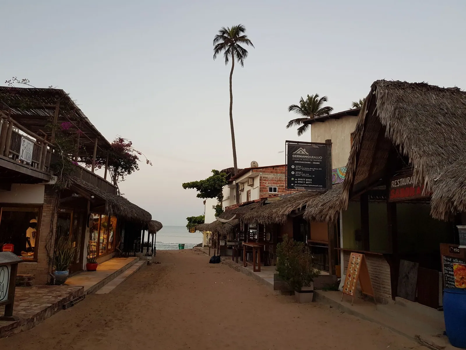 A sandy, palm-lined street of thatched shopfronts in Jericoacoara at dusk