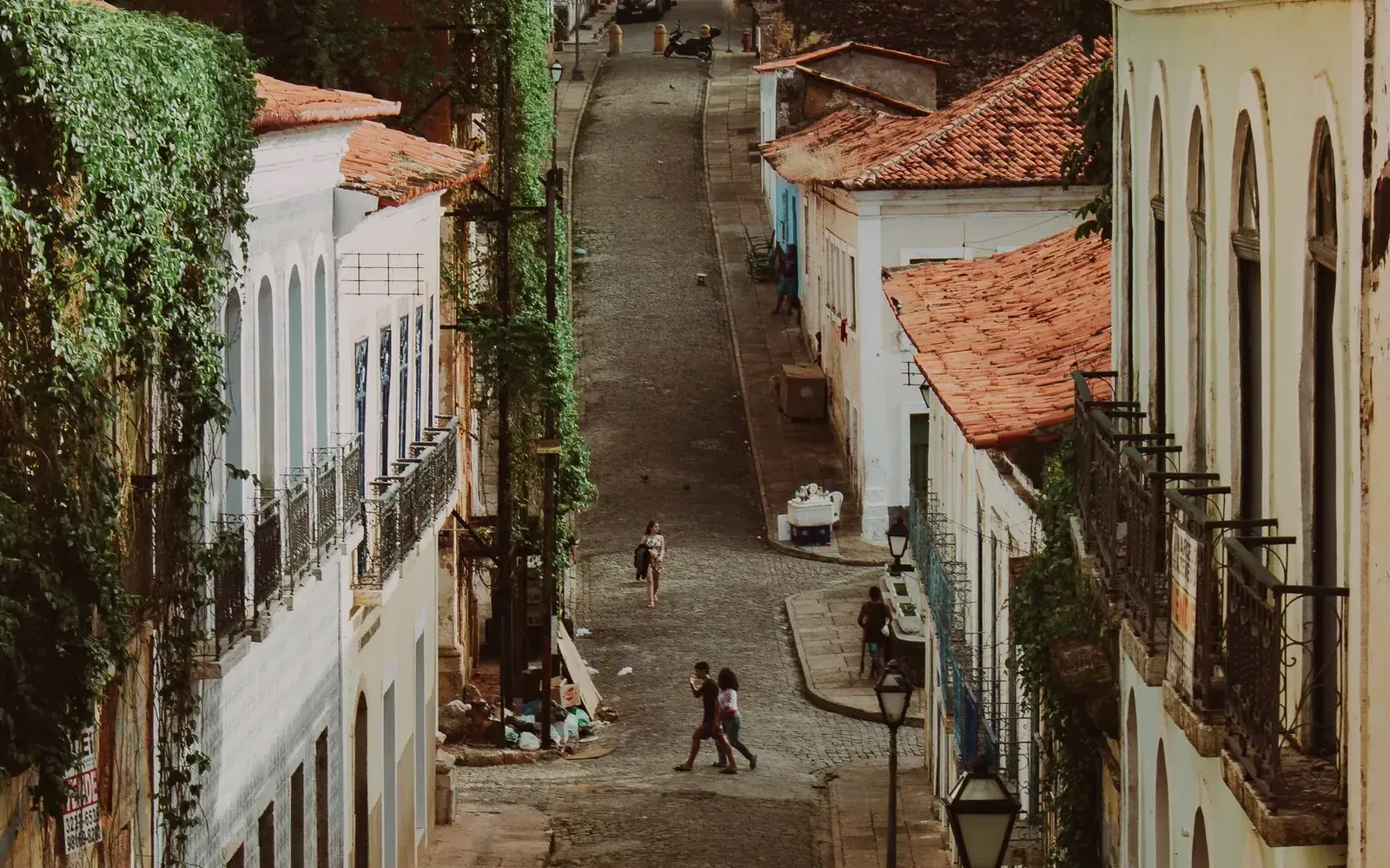 A steep colonial street in São Luís seen from above, weathered rooftops falling away below