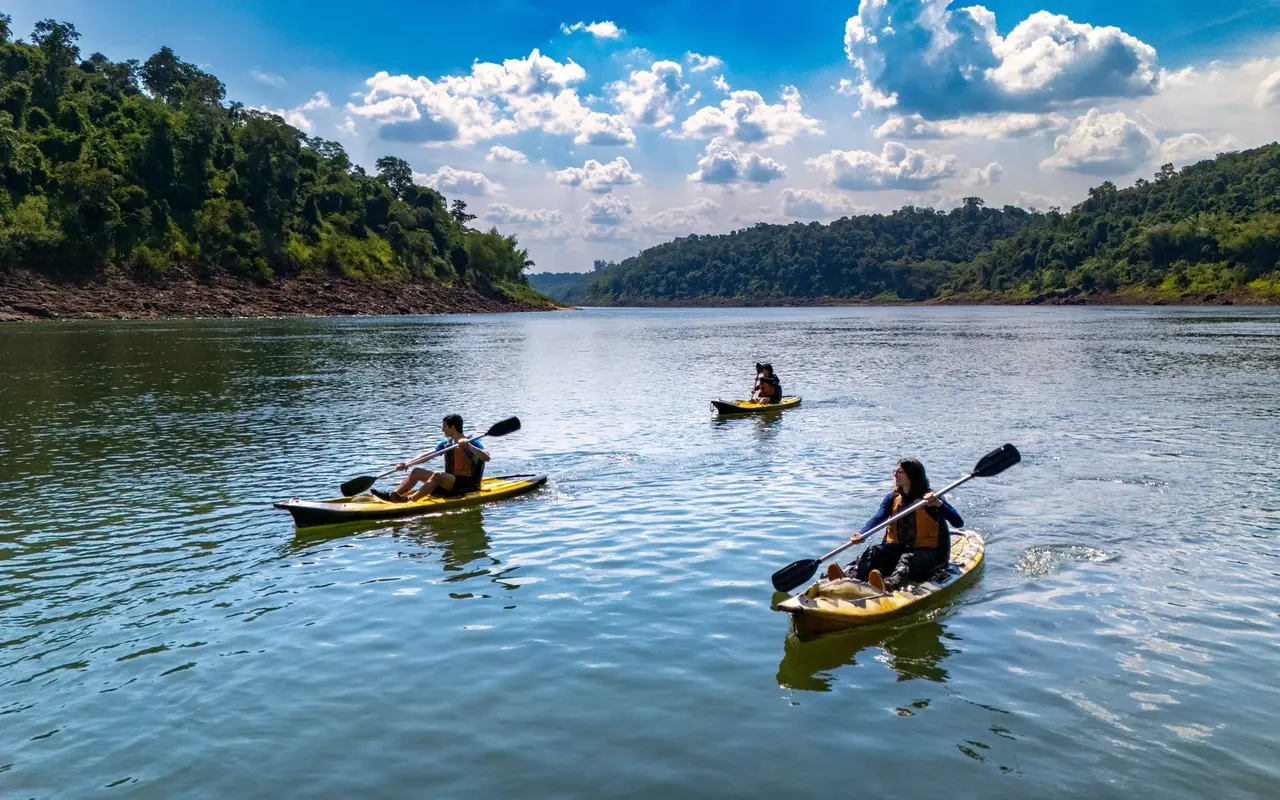 Kayakers paddling the wide calm Iguaçu River between forested banks under a blue sky