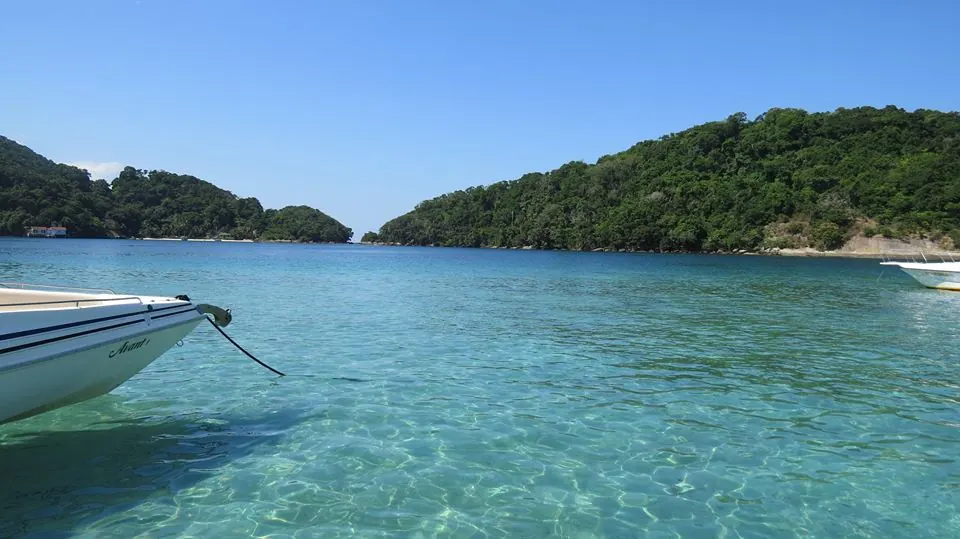 The bow of a boat over glass-clear turquoise water in a forested cove off Ilha Grande